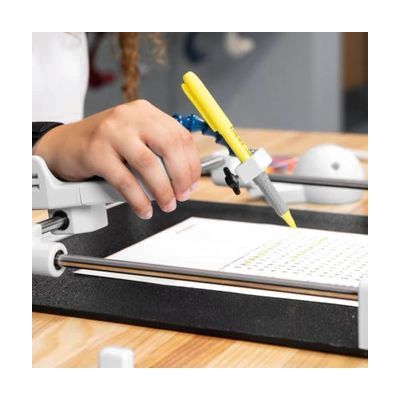 A writing-assistance device with a white armrest and clamp holding a yellow pen, used by a child's hand over a desk guide.