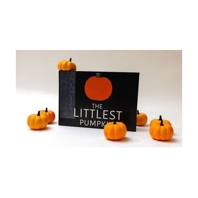 The Littlest Pumpkin storybook stood up on its long edge showing its cover with the title and a simple illustration of an orange pumpkin. Small decorative pumpkins are positioned around and on top of the book.