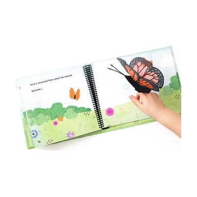 The hand of a young child is playing with a Monarch butterfly manipulative included in On the Way to Literacy: The Caterpillar. The book is opened to a page with bright and colorful illustrations of a butterfly flying over a field.
