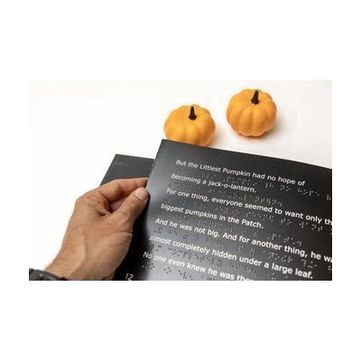 A man's hand holding the left edge of an open page with white large print text and UEB braille. Above the book are two small decorative pumpkins.