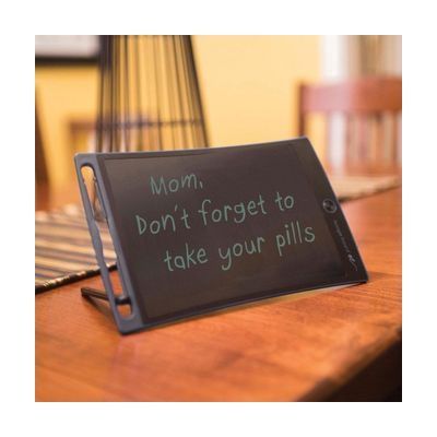 A black rectangular LCD writing tablet on a stand, propped on a wooden table with a handwritten reminder message.